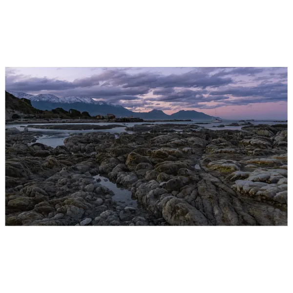 Long exposure photo of Jimmy Armers Beach, Kaikoura, with flat rocks in the foreground and snow-capped mountains in the distance.