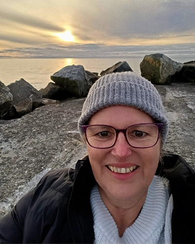 Jacqui Stokes bundled up in a woolly hat, jumper, and jacket, standing against a backdrop of a wintery sunrise over the sea.