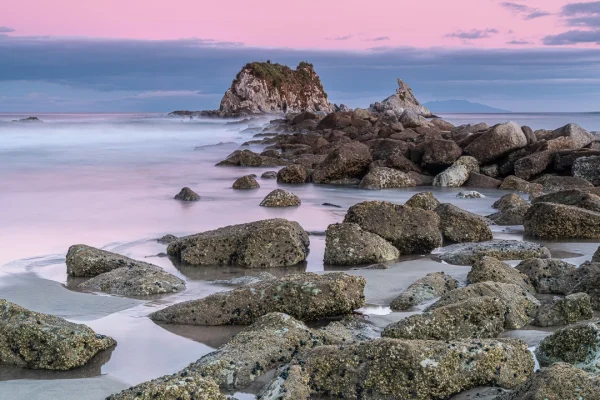 A leading line of rocks on Mangawhai Heads Beach guides the eye toward Sentinel Rock, with distant islands on the horizon and a soft pink-toned sky above